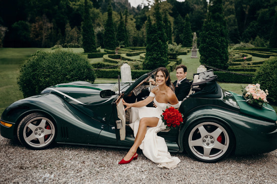 Bride and groom in sports car, bride is wearing red shoes and holding a bouquet of red roses